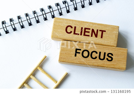 Client focus text written on a spiral notepad placed beside wooden blocks and ladder illustrating customer prioritization. 131168195