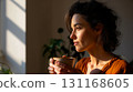 Soft sunlight frames woman with coffee; coffee warms hands as woman stands near window, calm profile and warm tone suggest mindful pause, coffee moment inspires cozy start and slow living theme 131168605