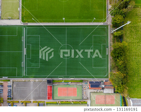 Aerial view of a sports complex with a full size soccer field, smaller fields, red surfaced tennis courts, surrounding buildings, parking areas, and tree lined boundaries. 131169191