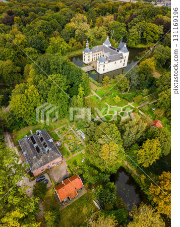 Aerial view of a white multi tower castle on a moat island, with geometric gardens, red roofed and brick buildings nearby, all surrounded by lush mixed forest in a serene heritage landscape. Aerial view of a white multi tower castle on a moat island, with geometric gardens, red roofed and brick buildings nearby, all surrounded by lush mixed forest in a serene heritage landscape. 131169196