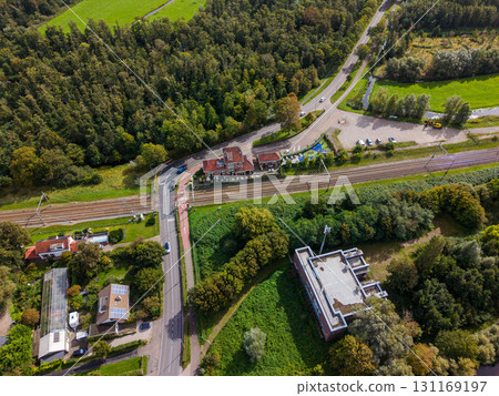 Aerial view of a semi rural area with a railway line, small station, parked cars, level crossing, wooded areas, open fields, and scattered buildings including one with distinctive architecture. 131169197