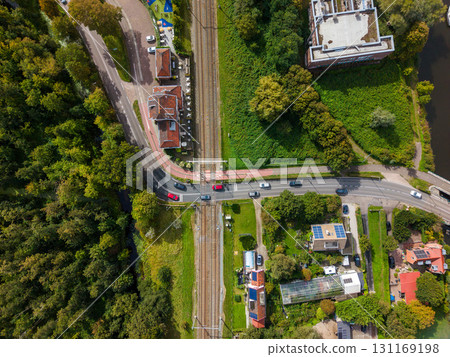 Aerial view of a semi urban area with a vertical railway line, road crossing with cars halted at barriers, red tiled and flat roof buildings, dense trees, and a canal near a large building. 131169198