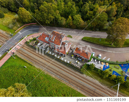 Aerial view of a red roofed cafe beside railway tracks and a road crossing with safety barriers, set in a green semi rural landscape of trees and vegetation. Aerial view of a red roofed cafe beside railway tracks and a road crossing with safety barriers, set in a green semi rural landscape of trees and vegetation. 131169199