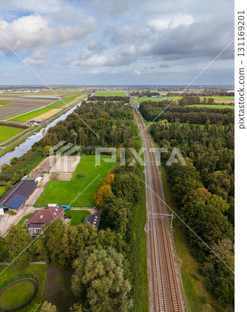 Aerial view of a straight railway flanked by dense trees, with fields, a canal, and solar roofed houses to the left, under a partly cloudy sky casting soft shadows. 131169201