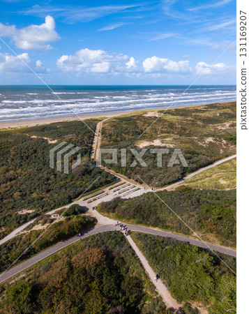 Aerial view of a bright coastal scene with a sandy beach, white capped waves, green dunes, and intersecting walking paths, dotted with people under a mostly clear sky. 131169207