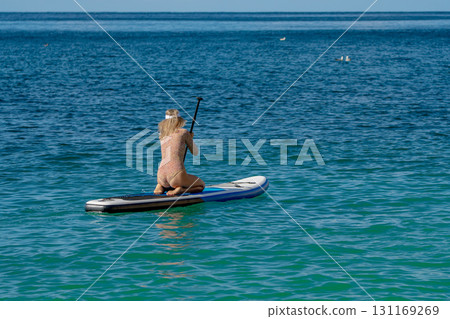 Woman Paddleboard Ocean: Kneeling female paddling for water sport on blue sea, summer recreation. Woman Paddleboard Ocean: Kneeling female paddling for water sport on blue sea, summer recreation. 131169269