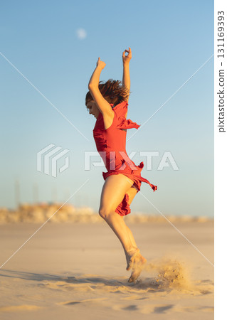 Dancer jumping on the beach at sunset wearing red dress 131169393