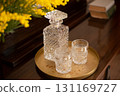 An elegant crystal decanter and two glasses sit on a vintage brass tray on a wooden table, next to a vase with yellow flowers and old books. 131169727
