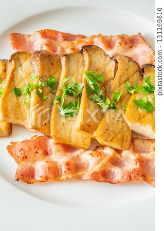 Fried eryngii mushrooms served with crispy bacon slices on white plate 131169810