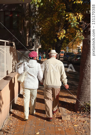 Elderly couple walking hand in hand on autumn street in warm sunlight, dressed in cozy outerwear and hats. Concept of senior love, retirement lifestyle, senior companionship 131170388