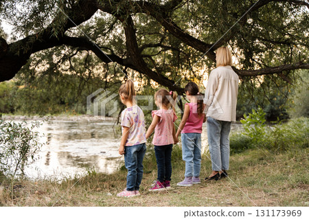 Mother and daughters holding hands looking at river under tree 131173969