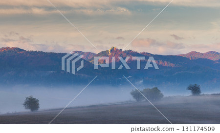 The Lietava castle in Slovakia, Europe. Autumn landscape with fog over fields and distant castle on hilltop at sunrise. Nature's scenic beauty. The Lietava castle in Slovakia, Europe. Autumn landscape with fog over fields and distant castle on hilltop at sunrise. Nature's scenic beauty. 131174509