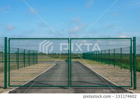 Secure metal gate with green fence enclosing private asphalt road leading to open rural landscape Secure metal gate with green fence enclosing private asphalt road leading to open rural landscape 131174602
