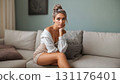 Half-length portrait of pretty thoughtful blonde caucasian young woman sitting on sofa and looking at camera in bright empty living room interior. Digital art illustration. 131176401