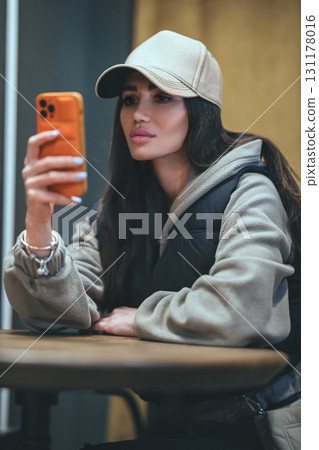 young woman with long dark hair sits comfortably at a wooden table wearing a cap and a cozy outfit. She gazes at her phone absorbed in what she sees enjoying a relaxed afternoon. young woman with long dark hair sits comfortably at a wooden table wearing a cap and a cozy outfit. She gazes at her phone absorbed in what she sees enjoying a relaxed afternoon. 131178016