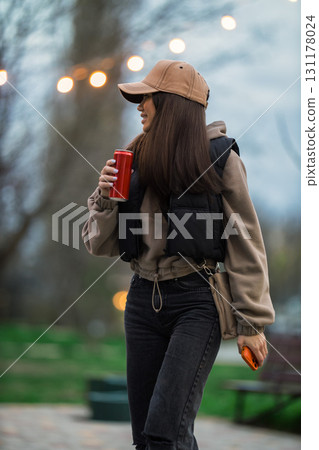 girl with a cap strolls through a park holding a red drink in one hand. She wears a stylish outfit and enjoys the warm ambiance of early evening lights. 131178024