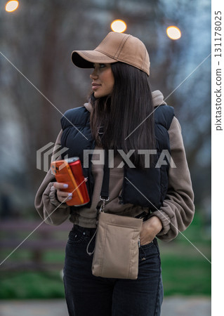 In a serene park setting during autumn a girl with long dark hair and a cap holds her phone. She is dressed in cozy attire enjoying her surroundings as she looks off thoughtfully. 131178025