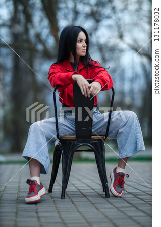 girl sits on a black chair with her legs apart wearing a bright red sweater and wide jeans. She is outdoors with trees and a cloudy sky in the background giving a relaxed vibe. 131178032