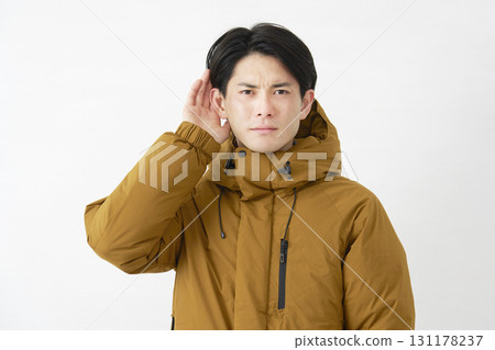 一位身穿羽絨外套的男子用手摀住耳朵聆聽，白色背景 131178237