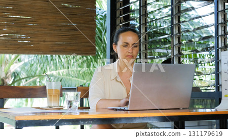Young woman sits on a terrace in a tropical setting focused on her laptop and enjoying cold coffee  131179619