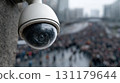Close up view of a white security dome camera with rain droplets on its round lens watching a blurred large multitude of people moving on a wide city street in urban setting. 131179644