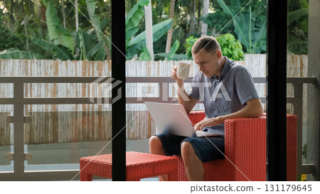 Man enjoys morning coffee while working on a laptop from a balcony surrounded by a lush tropical landscape  131179645
