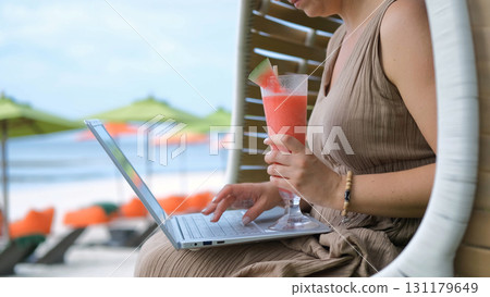 Close-up of young woman working on laptop in a woven chair sipping a watermelon shake while enjoying the ocean view  131179649