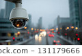 Automated security dome camera covered in rain droplets monitors a wet urban street with blurred moving vehicles. Bright traffic lights and car lights create a glowing distant view. 131179666