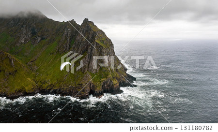 Paraglider flying over dramatic sea cliffs with rugged mountains and stormy ocean waves in ultra HD nature landscape 131180782