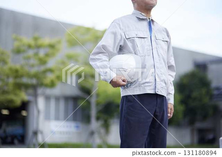 A senior man in work clothes stands outdoors, holding a helmet. A senior man in work clothes stands outdoors, holding a helmet. 131180894