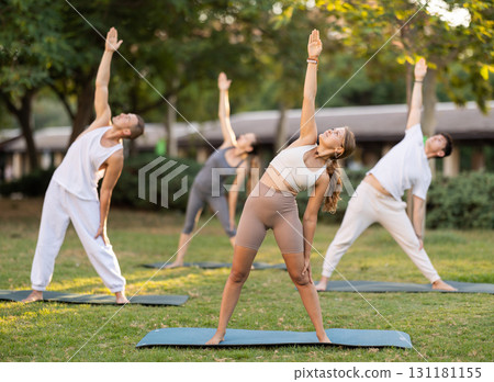 Yoga lesson visitors with coach perform Trikonasana in park 131181155