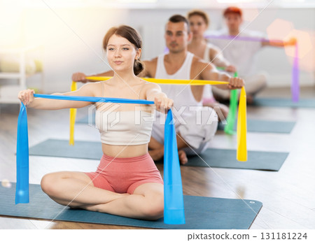Professional young girl wearing sports clothes using resistance bands during group pilates class sitting on mat in fitness room Professional young girl wearing sports clothes using resistance bands during group pilates class sitting on mat in fitness room 131181224