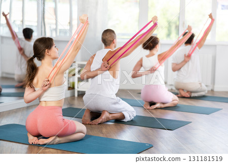 Young men and women doing shoulder workout with resistance band sitting on knees 131181519