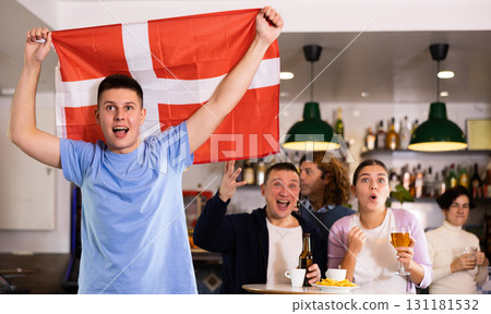 Company of young adult sports fans supporting Danish team with state flag while drinking beer in bar Company of young adult sports fans supporting Danish team with state flag while drinking beer in bar 131181532