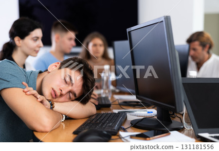 Exhausted guy student sits at table, puts head on crossed arms and sleeps after computer classes Exhausted guy student sits at table, puts head on crossed arms and sleeps after computer classes 131181556