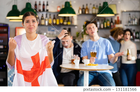Company of upset young adult sports fans holding flag of England and worrying about defeat during game match on TV in pub 131181566