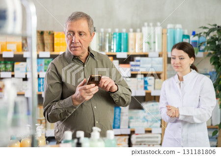 Male customer looking for something in drugstore, checking the list on her smartphone. Female pharmacist offers him medicines Male customer looking for something in drugstore, checking the list on her smartphone. Female pharmacist offers him medicines 131181806