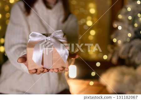 Gift, Christmas, Woman - Woman holding gift box with white ribbon in front of Christmas lights. 131182067
