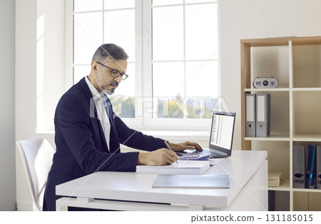 Male accountant or bookkeeper working in office, using laptop, and taking notes in accounting book 131185015