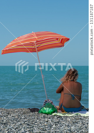 A girl in a blue swimsuit sits in the shade of a red umbrella on the seashore. A girl in a blue swimsuit sits in the shade of a red umbrella on the seashore. 131187187