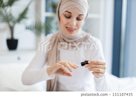 Muslim woman of young age, wearing neutral hijab, applying nail polish indoors with focus on self-care, beauty, relaxation. Soft natural light, bright modern interior, serene expression Muslim woman of young age, wearing neutral hijab, applying nail polish indoors with focus on self-care, beauty, relaxation. Soft natural light, bright modern interior, serene expression 131187435