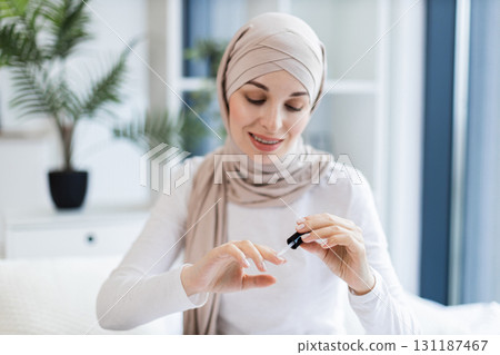 Young Muslim woman wearing head scarf applying nail polish while sitting indoors. Concept of self-care, beauty, and femininity in a modern home environment. Focus on hands and personal grooming. Young Muslim woman wearing head scarf applying nail polish while sitting indoors. Concept of self-care, beauty, and femininity in a modern home environment. Focus on hands and personal grooming. 131187467
