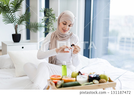 Muslim woman around twenties wearing hijab applies nail oil sitting indoors, surrounded by natural care items such as fruits and oils, portraying a health-conscious lifestyle on self-care and wellness 131187472