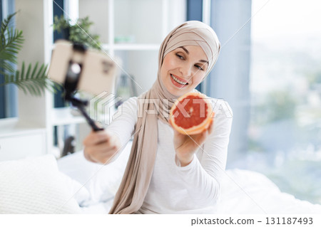 Young Muslim woman wearing hijab filming video blog holding grapefruit for skincare routine in bright room. Promoting natural cosmetic ingredients. Young Muslim woman wearing hijab filming video blog holding grapefruit for skincare routine in bright room. Promoting natural cosmetic ingredients. 131187493