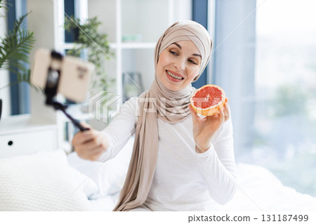 Muslim woman wearing hijab holding grapefruit recording video blog in home interior. Showcasing creative concepts and healthy lifestyle in cheerful atmosphere. Promoting natural skincare ideas. 131187499