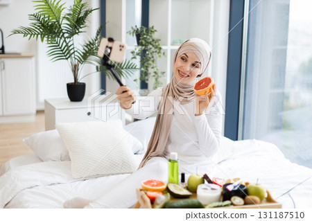 Muslim adult woman in white hijab filming beauty blog on phone about grapefruit benefits for natural facial skin care. Cozy home environment with natural light and healthy organic fruits. 131187510