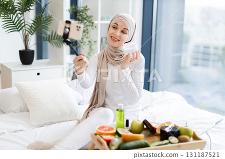 Adult Muslim woman wearing hijab filming content using phone about facial cream and skincare. Tray with fruits and cosmetics visible on bed in modern, bright room. Adult Muslim woman wearing hijab filming content using phone about facial cream and skincare. Tray with fruits and cosmetics visible on bed in modern, bright room. 131187521