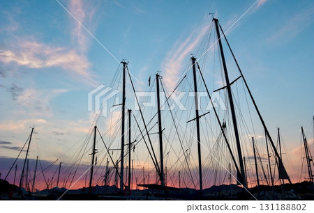 A tranquil harbor at sunset with colorful clouds and a vibrant red-orange sky reflecting on the calm water, framed by silhouettes of docked sailboats. 131188802