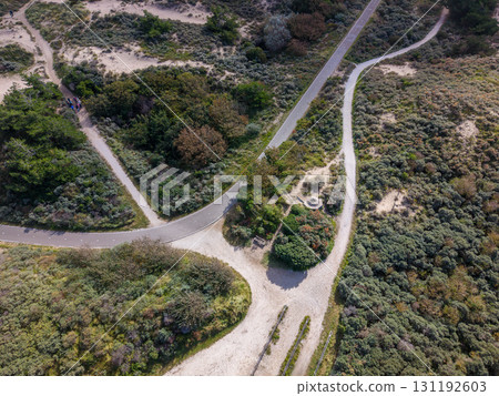 Aerial view of winding paved and dirt paths weaving through green shrubs, sandy terrain, and trees, with a circular stone feature nestled among the vegetation. 131192603