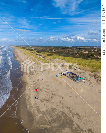 Aerial view of a sandy beach bordered by vegetated dunes, with a small building nearby, people on the shore, and colorful kites hinting at kite surfing under a partly cloudy sky. 131192604